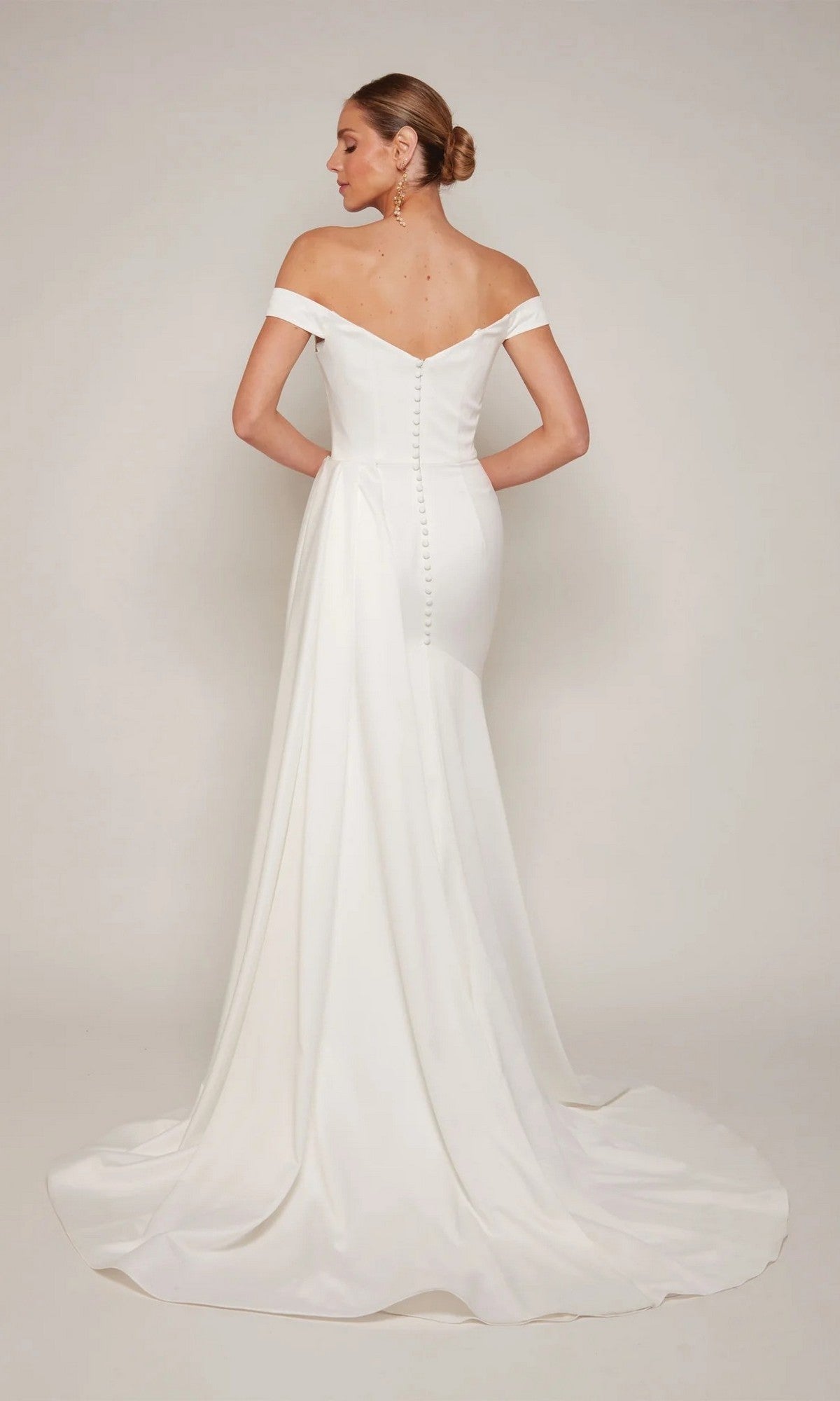 A woman with her hair in a low bun faces away, wearing the Alyce 7098 long formal dress—an off-the-shoulder white gown featuring a row of buttons down the back and a sweeping train.